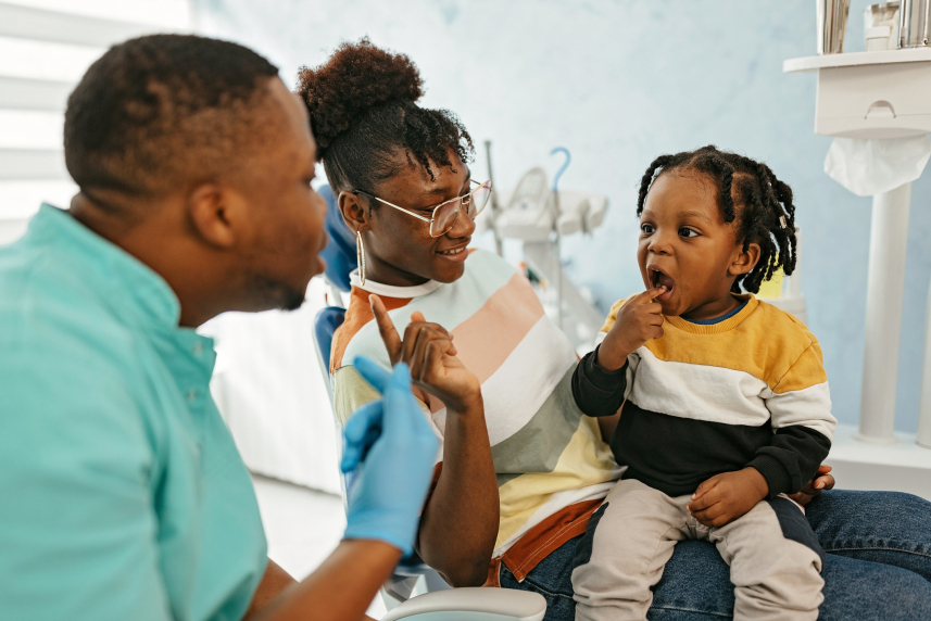 A child sits on an adult's lap next to a dentist.